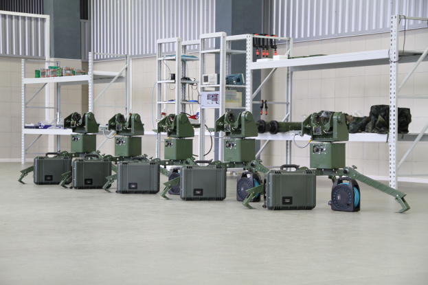 Eine Gruppe von Militärrobotern auf einem Boden, umgeben von Koffern und anderen Gegenständen auf Gestellen, mit einer Wand und einem Geländer im Hintergrund.