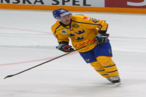 Ein Hockey-Spieler in Aktion auf dem Eis, der einen Helm, Handschuhe und Schlittschuhe trägt und einen Hockey-Schläger hält, mit einer Tafel mit Text im Hintergrund.