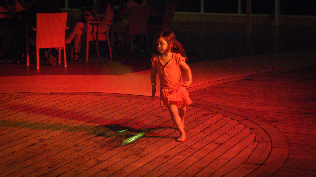 Ein Kind läuft auf einer hölzernen Plattform drinnen, mit einigen Menschen im Hintergrund auf Stühlen sitzend.