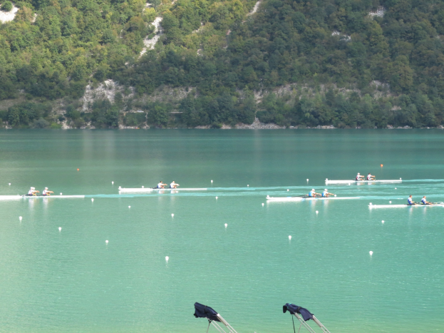 Eine Gruppe von Menschen, die in einem Wettbewerb auf einem Gewässer mit Bäumen und einem Hügel im Hintergrund rudern, mit Kleidung, die über Stände am Boden drapiert ist.
