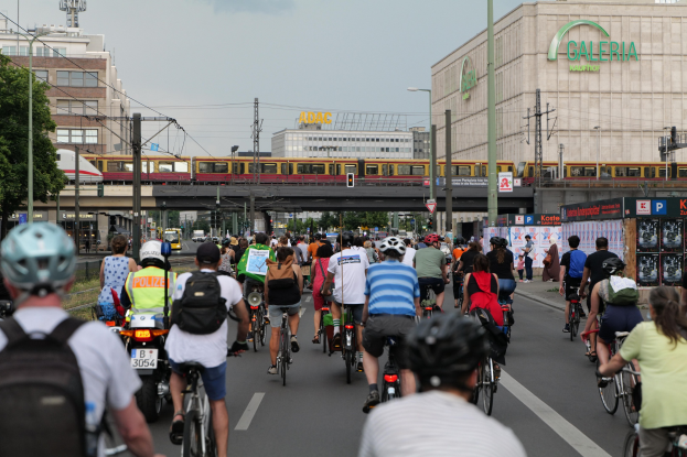 Eine Gruppe von Menschen, die auf Fahrrädern eine Straße mit hohen Gebäuden entlangfahren, einige tragen Helme und Taschen, mit einem Zug, Strommasten, Bäumen und einem klaren blauen Himmel im Hintergrund.