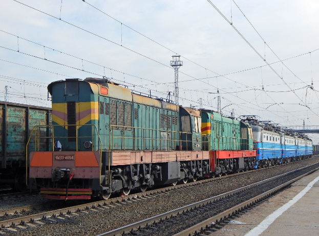 Diesellokomotive fährt auf Schienen neben einem Bahnsteig mit elektrischer Infrastruktur und einem klaren Himmel.