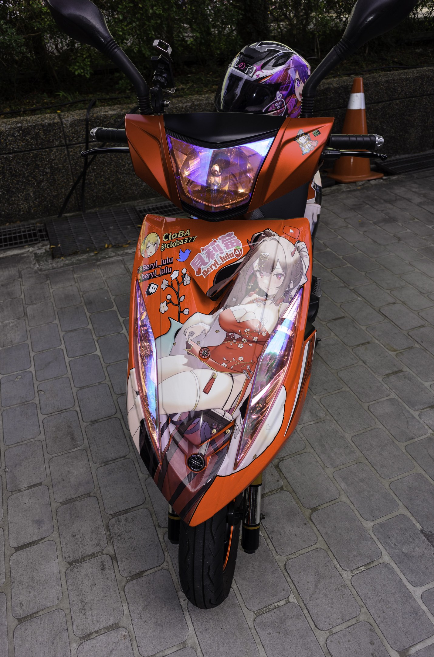 Ein oranger Roller auf einer Straße mit einem Helm daneben, neben einem Verkehrskegel, einer Wand und Bäumen im Hintergrund.