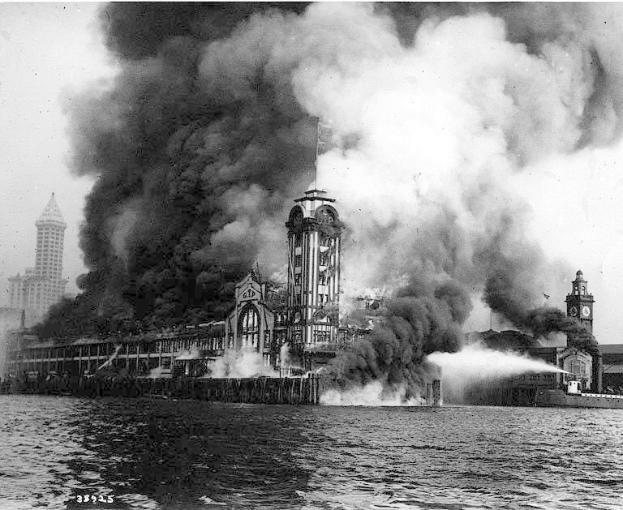 Schwarz-weiß-Foto eines großen, von Flammen eingehüllten Gebäudes mit Rauch, der daraus aufsteigt, einem Gewässer im Vordergrund und anderen Gebäuden und Himmel im Hintergrund.