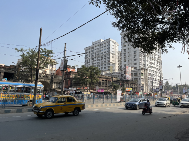 Gelbes Taxi auf belebter Straße in Mumbai unterwegs, umgeben von hohen Gebäuden, Bäumen, Strommasten mit Drähten, Laternen und Schildern unter einem sichtbaren Himmel.