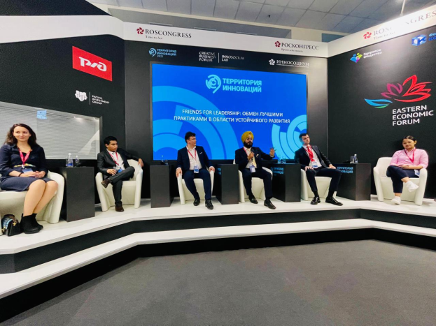 Eine Gruppe von Menschen sitzt auf einer Bühne beim Östlichen Wirtschaftsforum in Moskau, mit Flaschen und anderen Gegenständen auf den Tischen, Banner mit Text und Logos im Hintergrund und Deckenbeleuchtung.