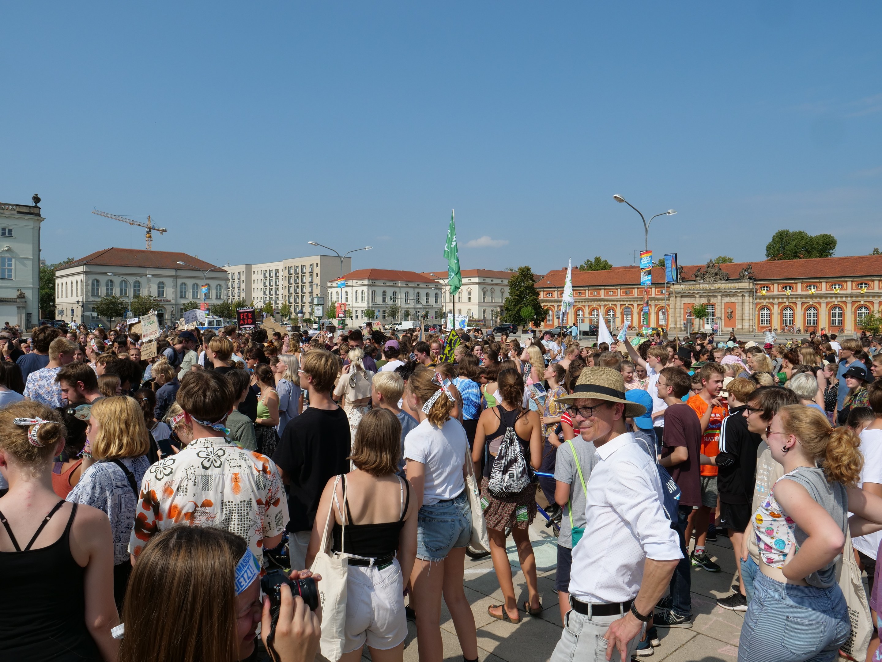 Große Menschenmenge vor einem Gebäude mit Flaggen, Bäumen und Laternen im Hintergrund, einige halten Kameras während einer Demonstration.