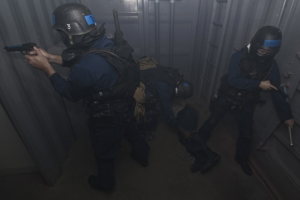 Eine Gruppe von Polizisten in Einsatzausrüstung, einer hält eine Waffe, steht neben einem Metallcontainer und einer Leiter in einem begrenzten Raum.