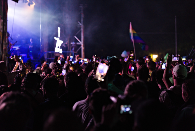Eine Menge bei einem Konzert hält leuchtende Handys hoch, während eine kleine Band auf der Bühne unter hellen Lichtern spielt.