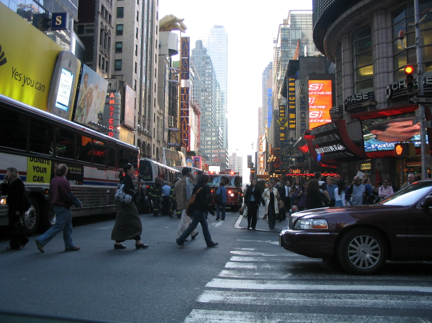 Eine belebte Stadtstraße mit Fahrzeugen, Fußgängern, die eine Zebrastreifen überqueren, Gebäuden, Schildern, Verkehrsampeln und einem klaren blauen Himmel.