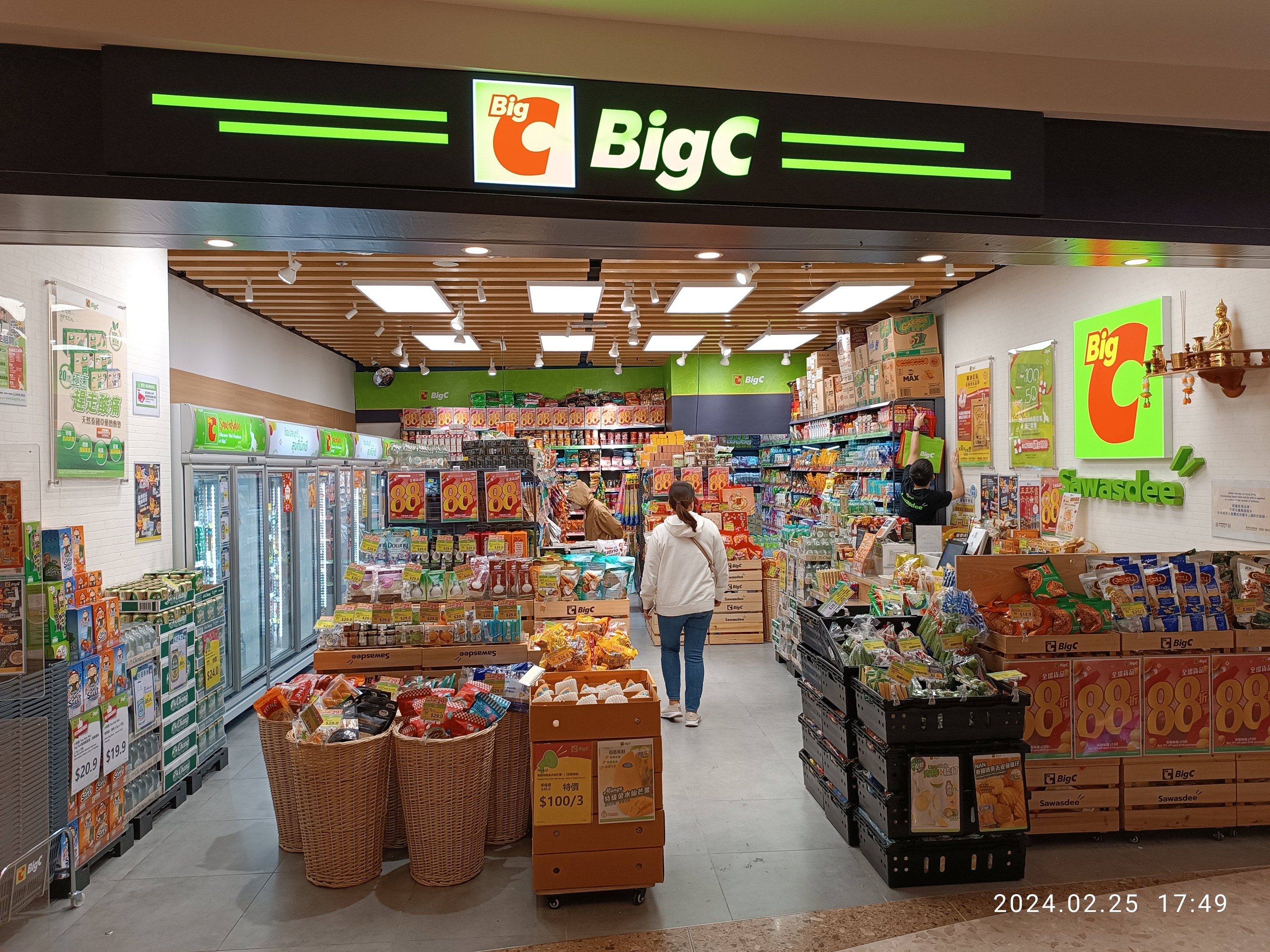 Eine Person in einem weiß-blauen Kleid steht vor einem hell erleuchteten Supermarktgang mit verschiedenen Lebensmitteln, während Kölzer, Wandtafeln und Deckenleuchten im Hintergrund zu sehen sind.