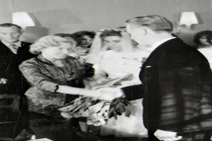 Ein Schwarz-Weiß-Foto von einer Braut und einem Bräutigam, die sich Ringe anstecken, wobei die Braut ein weißes Kleid und eine Schleppe trägt und einen Strauß hält und der Bräutigam neben ihr steht und einen Anzug und eine Krawatte trägt, mit Wandlampen im Hintergrund.