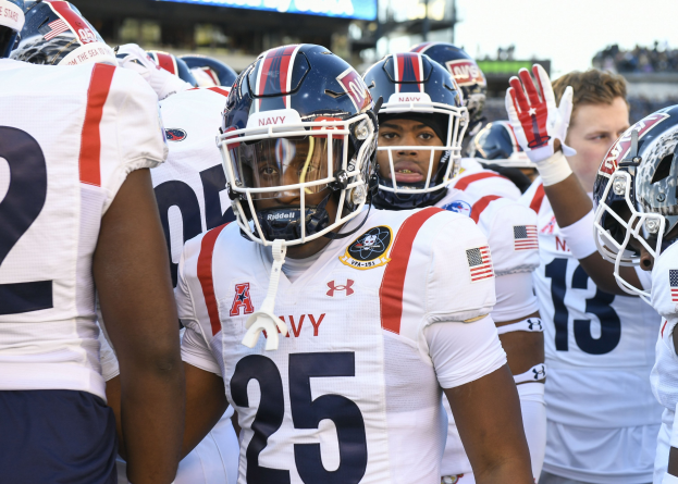 Gruppe von Männern in Football-Uniformen auf einem Feld mit Helmen und Handschuhen, mit einer unscharfen Menge, einem Scoreboard mit 'Auburn vs Virginia Tech - 11/28/15 College Football Pick, Odds, and Prediction' und Himmel im Hintergrund.