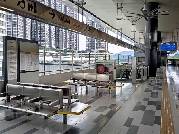 Ein leerer Flughafen Wartebereich mit zahlreichen Bänken, Informationsanzeigen, Säulen, einem Deckenventilator und Lampen, mit Gebäuden, Bäumen, Wasser und einem klaren blauen Himmel im Hintergrund.