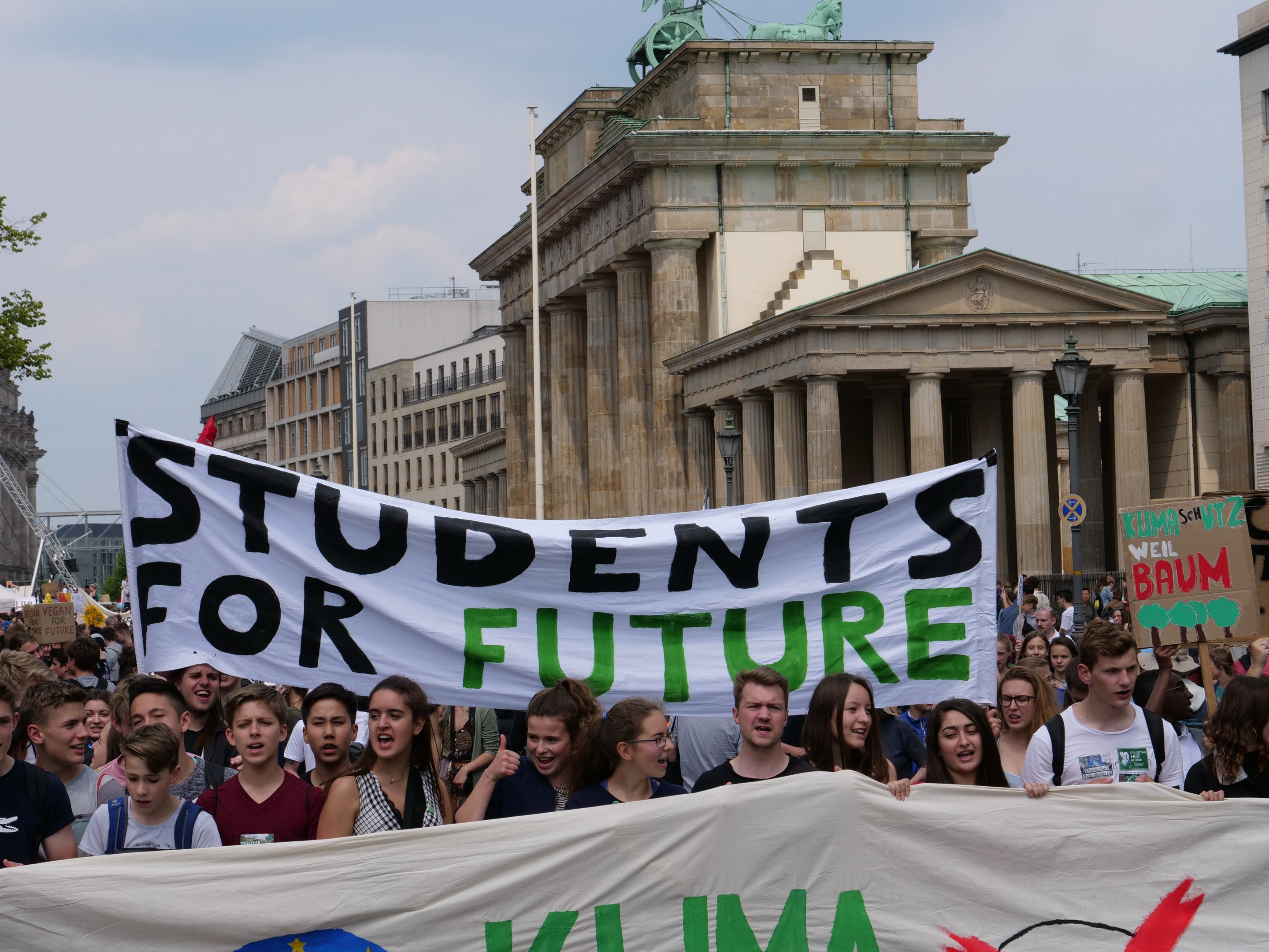 Gruppe von Studenten marschiert in Berlin mit einem bunt bemalten "Students for Future"-Schild an Gebäuden, Bäumen und Himmel vorbei.
