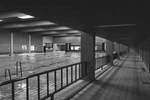 Ein leerer Swimmingpool in einem verlassenen Gebäude, mit einem gefliesten Boden, Geländern, Säulen, Wänden, Fenstern und Deckenlampen, in Schwarz-Weiß aufgenommen.