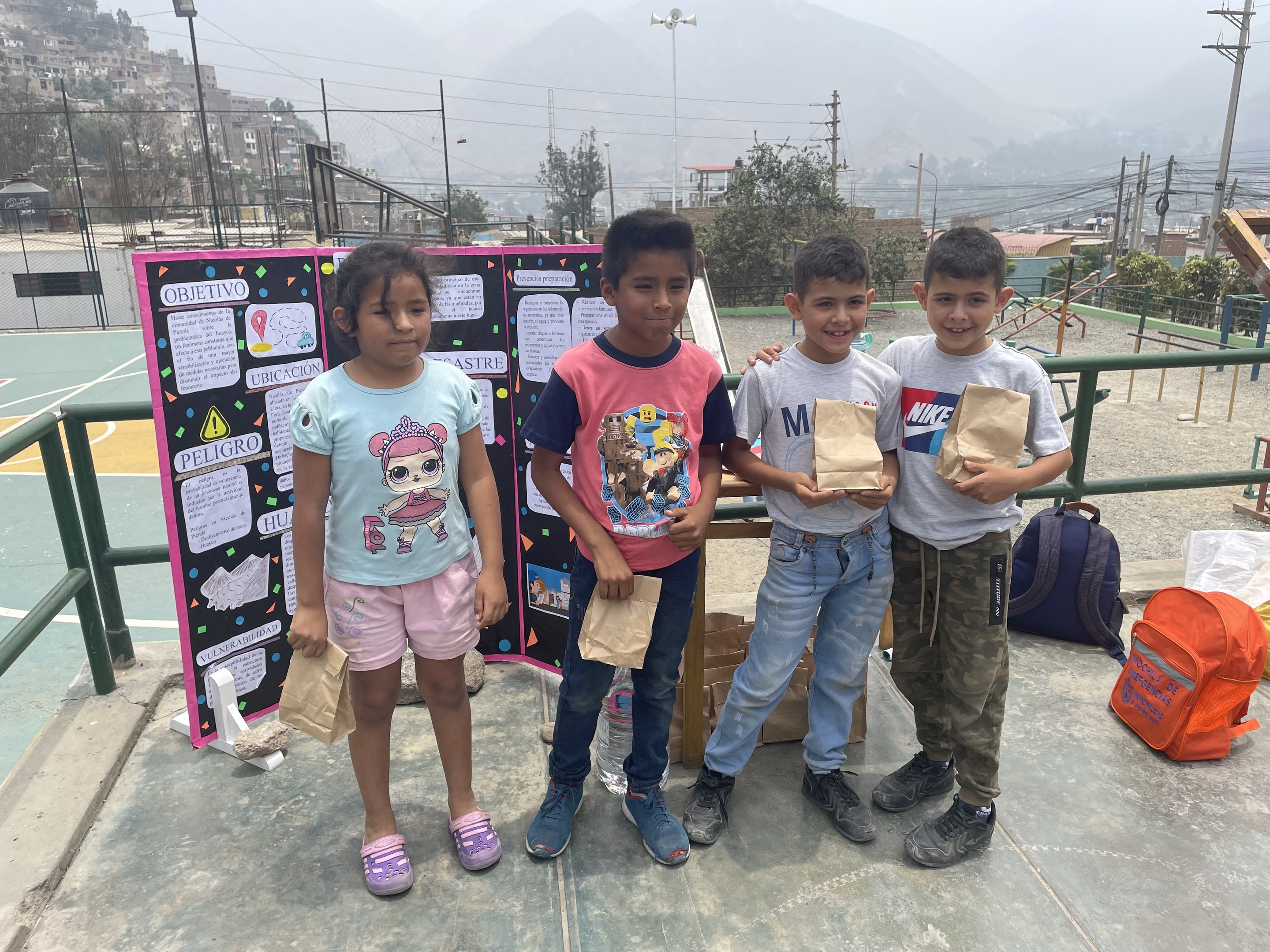 Gruppe von Kindern, die Papiertüten auf einem Basketballfeld halten, mit Vorräten auf dem Boden und einer Tafel mit Text links daneben, vor dem Hintergrund von Zäunen, Pfosten, Bäumen, Gebäuden, Hügeln und Himmel.