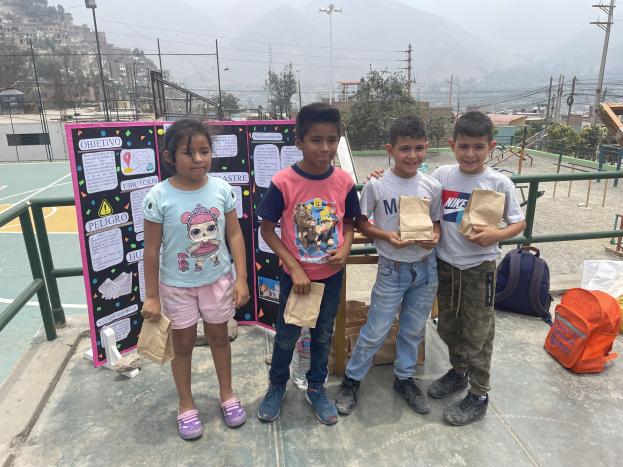 Gruppe von Kindern, die Papiertüten auf einem Basketballfeld halten, mit Vorräten auf dem Boden und einer Tafel mit Text links daneben, vor dem Hintergrund von Zäunen, Pfosten, Bäumen, Gebäuden, Hügeln und Himmel.