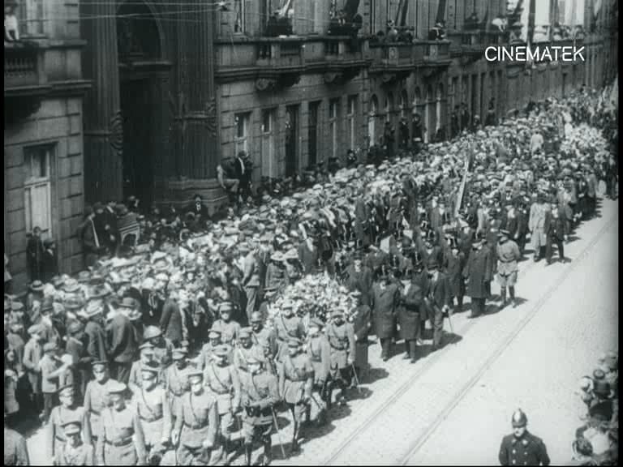 Schwarzes und weißes Foto einer großen Menge, die eine Straße entlangmarschiert, einige halten Gewehre, vor einem Gebäude, mit einem Wasserzeichen oben rechts.