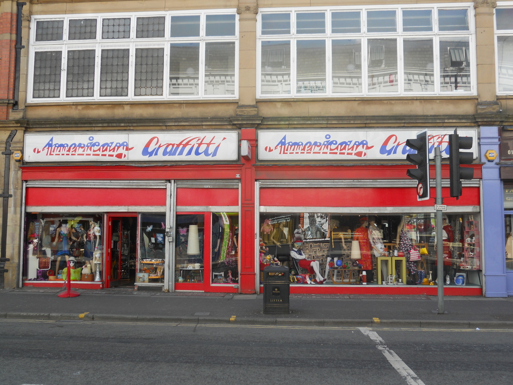 Eine Straßenszene mit einem Gebäude, Schildern, Puppen, Gläsern, einem Müllcontainer, einem Pfahl und Verkehrsampeln.