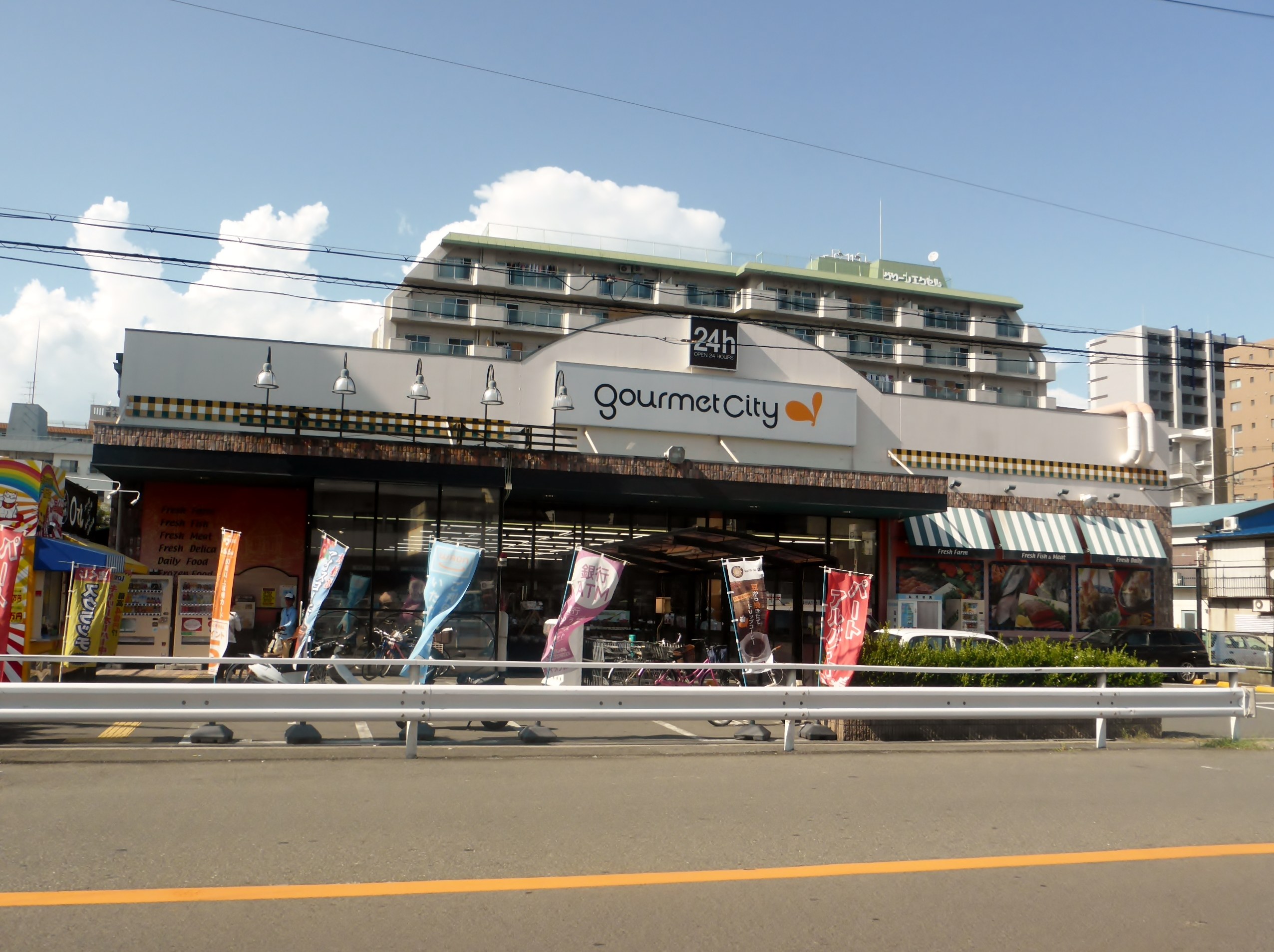 Eine belebte Stadtstraße mit Gebäuden, Straßenlaternen, Werbetafeln, Fahrzeugen, Absperrpollern, Büschen, Bäumen und einem bewölkten Himmel, mit einem Gourmetrestaurant in der Mitte.