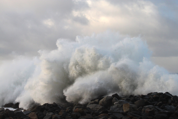 Eine große Welle schlägt gegen Felsen am Strand, mit Wasser, das gegen die Küste schwappt und einem bewölkten Himmel im Hintergrund.