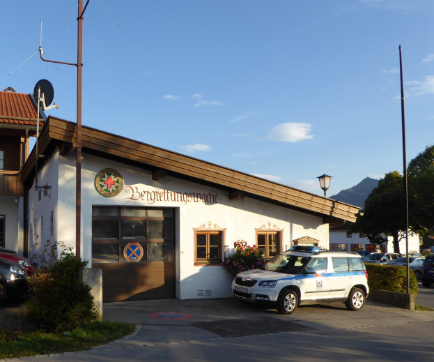Polizeiauto vor einem Gebäude mit Fenstern und Türen geparkt, umgeben von Grünflächen einschließlich blühender Pflanzen, Gras, Bäumen, Straßenmasten, einer Schüsselantenne und einem Schild unter einem bewölkten Himmel.