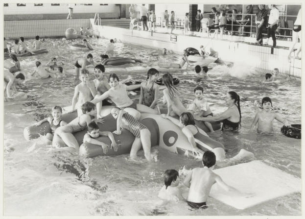 Schwarzes und weißes Bild von Kindern, die in einem Swimmingpool spielen, einige sitzen auf aufblasbaren Gegenständen und andere stehen, mit einer Wand und einem Geländer im Hintergrund.