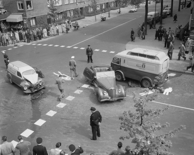 Schwarz-weißes Bild eines Autounfalls an einer Straßenkreuzung mit einem Van, anderen Fahrzeugen und einer Gruppe von Menschen, mit Gebäuden, Bäumen, Pfählen und Texttafeln im Hintergrund.