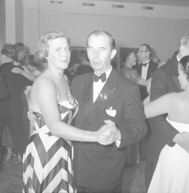 Ein Mann in einem schwarzen Blazer, weißem Hemd und schwarzen Hosen tanzt mit einer Frau in einem unbekannten Outfit auf einer formellen Veranstaltung, indem sie sich an den Händen halten und zur Musik schwenken, nahe einer Wand.