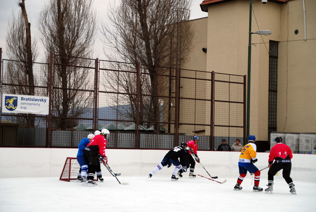 Menschen spielen Eisockey auf einer Eisfläche mit Gebäuden, Bäumen, einer Straßenlaterne, einem Namensschild und Zäunen im Hintergrund unter einem klaren Himmel.