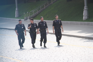 Drei Polizeibeamte in Uniform und Mützen gehen eine Straße mit Pfählen, Geländern, Treppen und Gras im Hintergrund entlang.