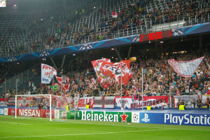 Ein Fussballspiel wird in einem hell erleuchteten Stadion mit einer grossen Zuschauermenge, einem Tor in der Mitte, Bannern mit Text und Logos drum herum und einem Bildschirm in der oberen linken Ecke gespielt.