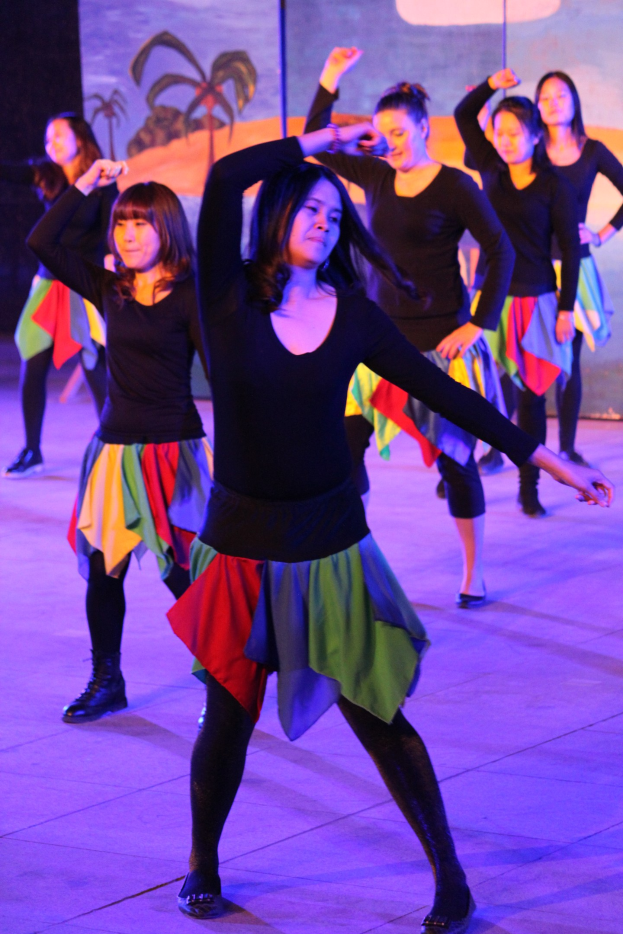 Junge Frauen in bunten Röcken und Schuhen tanzen energisch auf einer Bühne mit einem Bild im Hintergrund.