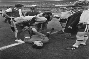 Schwarzes und weißes Bild eines Fußballspielers, der auf dem Boden liegt und von einer Gruppe Männer umgeben ist, mit einer Zuschauermenge im Stadionhintergrund.