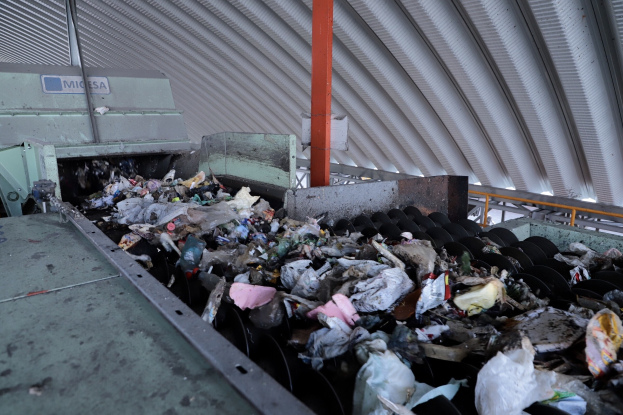 Großer Müllberg auf einem Förderband in einer Recycling-Anlage mit Maschinen und einem Metalldach.