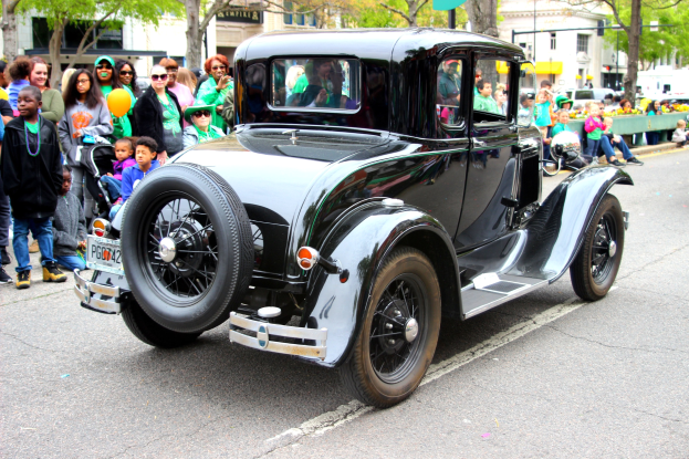 Ein Oldtimer fährt an einer Menge Menschen vorbei, die Hüte und Schutzbrillen tragen, mit Bäumen, Gebäuden, Laternenmasten und Texttafeln im Hintergrund.