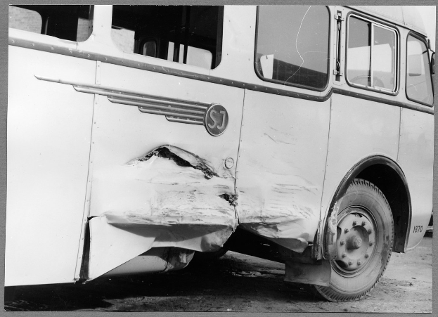 Schwarzes und weißes Foto eines verunglückten Busses mit eingedellter Front, zersplitterten Fenstern und geöffneter Fahrertür, die den Innenraum zeigt.
