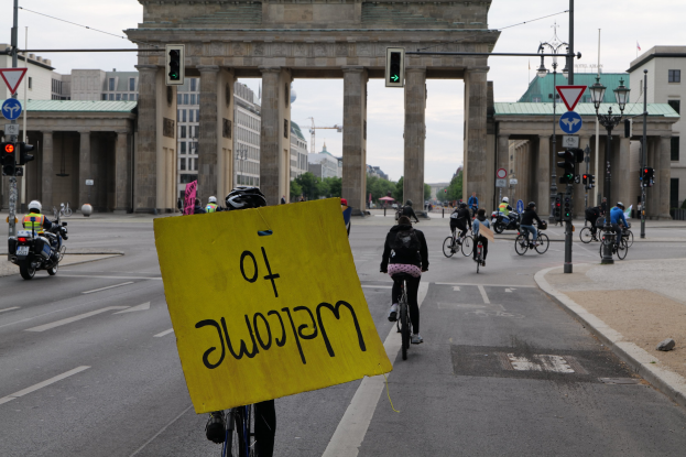 Eine Gruppe von Radfahrern fährt an der Brandenburgertor in Berlin, Deutschland, vorbei, einer hält ein gelbes Schild; die Straße ist mit Masten und Signalen gesäumt unter einem klaren Himmel.