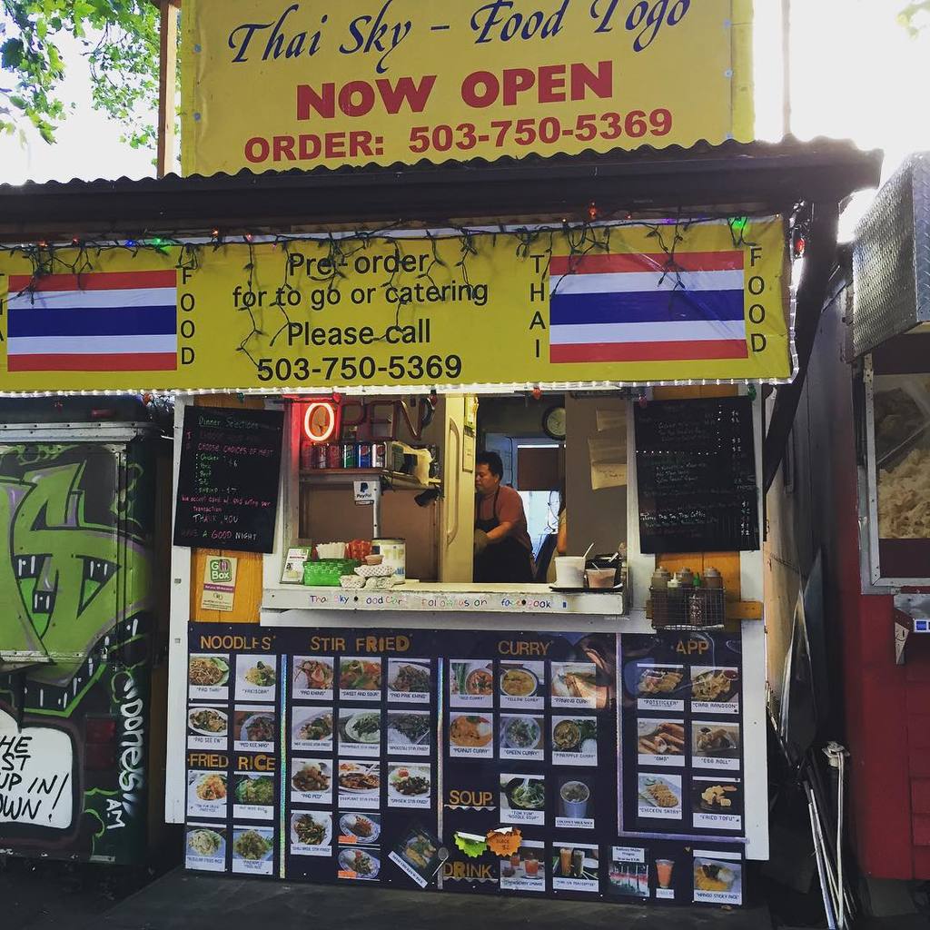 Ein Geschäft mit verschiedenen Artikeln wie Getränkedosen, Speisekarten, einer Uhr, Gewürzen in Plastikbehältern, Kondimenten, Werbetafeln und Namensschildern unter einem Himmel.