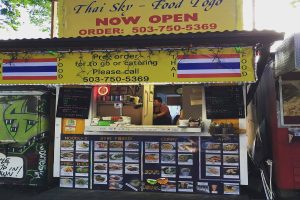 Ein Geschäft mit verschiedenen Artikeln wie Getränkedosen, Speisekarten, einer Uhr, Gewürzen in Plastikbehältern, Kondimenten, Werbetafeln und Namensschildern unter einem Himmel.