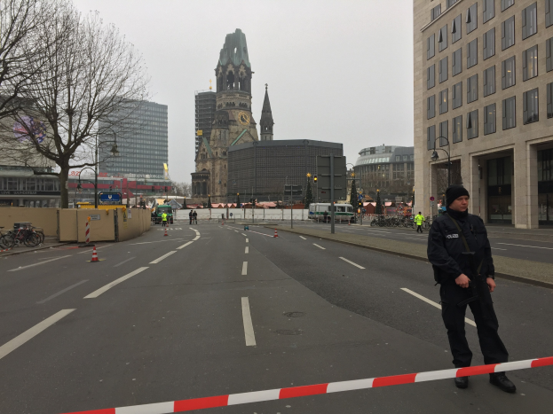Ein Polizist in schwarzer Uniform und Mütze steht auf einer Straße in Berlin umgeben von Verkehrskegeln, Fahrrädern, Laternenmasten, Bäumen und Gebäuden, mit einem Uhrenturm und blauem Himmel im Hintergrund.