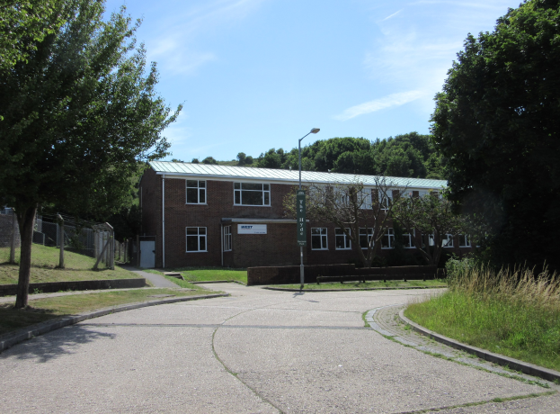 Ein Schulgebäude mit Fenstern, einer Straßenlaterne, einem Schild, einer Baumgruppe, Gras, einem Zaun und einem bewölkten Himmel, das inmitten eines Parks umgeben von saftigem Grün liegt.