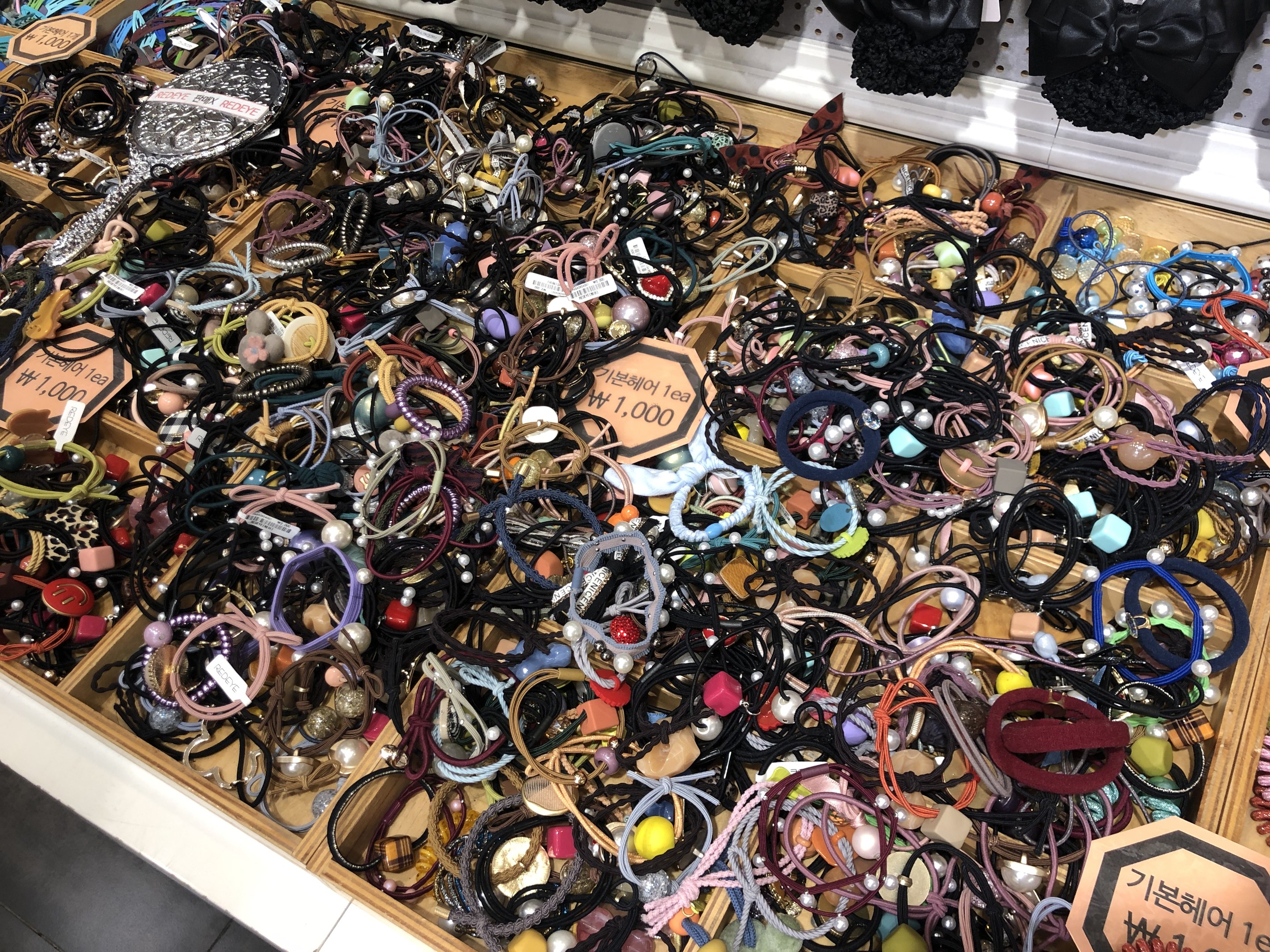 Vitrine mit verschiedenen Schmuckstücken einschließlich Armbändern, Ketten und Preisschildern neben hängenden Kleidungsstücken in einem Marktumfeld.