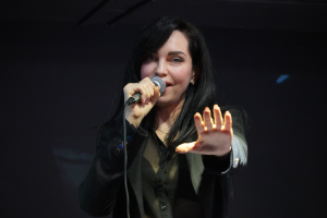 Eine Frau in einem schwarzen Kleid singt in ein Mikrofon auf einer Bühne, ihre Augen geschlossen und ihr Gesichtsausdruck voller Emotion und Konzentration, beleuchtet von einem hellen Licht.