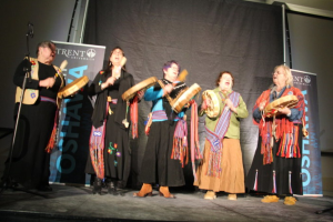 Eine Gruppe von Frauen in bunten Kleidern, die singen und Trommeln halten, mit einem schwarzen Vorhang und zwei blauen Rollplakaten dahinter.