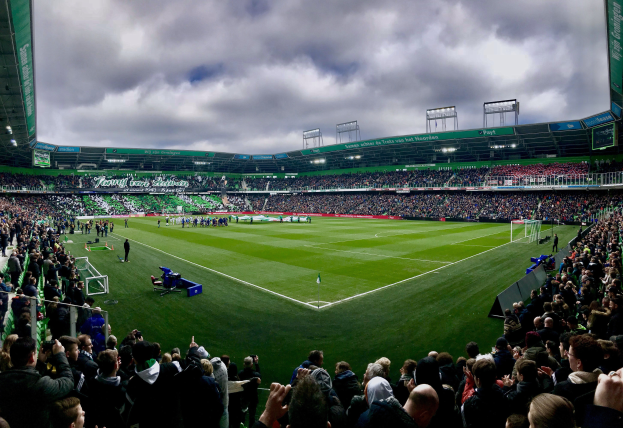 Eine große Menschenmenge in einem Stadion, die ein Fußballspiel schaut, wobei einige stehen und einige auf den Tribünen sitzen und verschiedene Gegenstände auf dem Feld unter einem bewölkten Himmel verstreut sind.