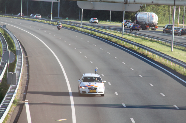 Ein Auto fährt auf einer Autobahn mit einem Polizeiwagen am Straßenrand, anderen Fahrzeugen, Leitplanken, Polen, Schildern und Bäumen im Hintergrund.