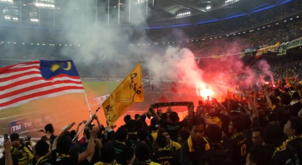 Eine große Menschenmenge in einem Stadion mit brennenden Fackeln in der Luft, einige halten Fahnen und eine Person hält eine Kamera, mit Rauch im Hintergrund und Schildern mit der Aufschrift "Malaysia vs Malaysia Live Streaming".