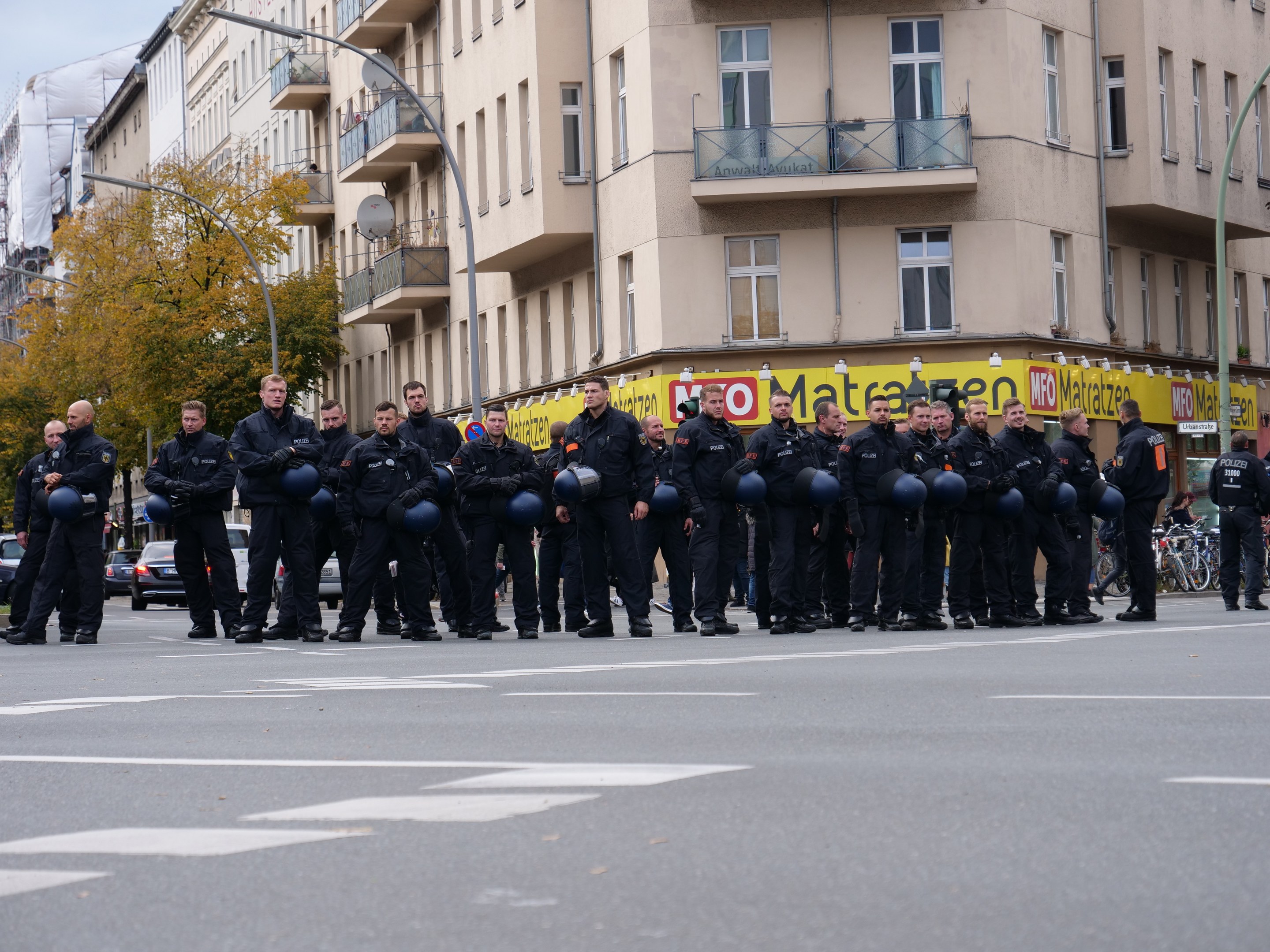 Gruppe von Polizisten in schwarzen Uniformen mit blauen Helmen, die auf einer Straße mit Laternen, Bäumen und Glasfenstern unter einem klaren blauen Himmel stehen.